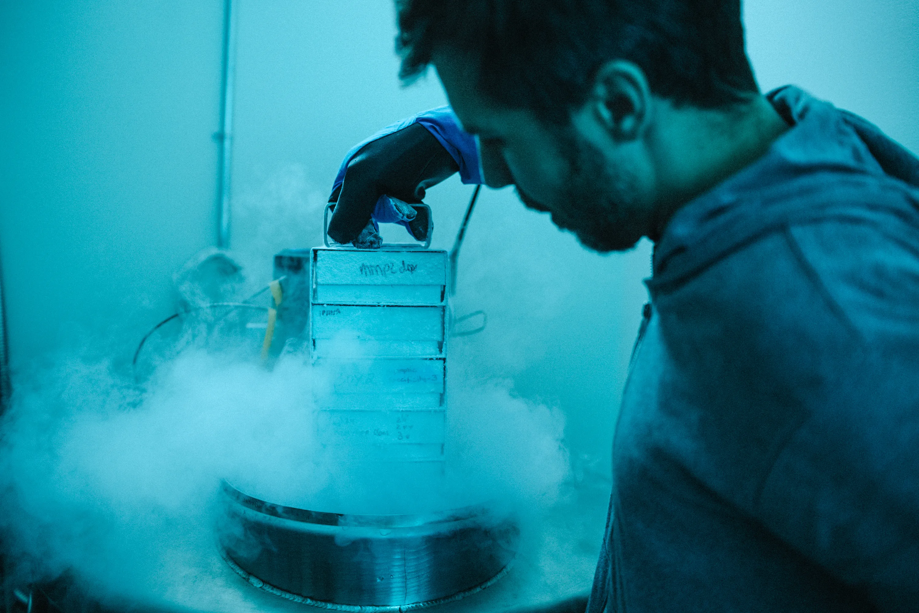 A photo of a scientist removing a vertical tray of boxes from a cold storage tank. Cold vapor rises from the open tank.