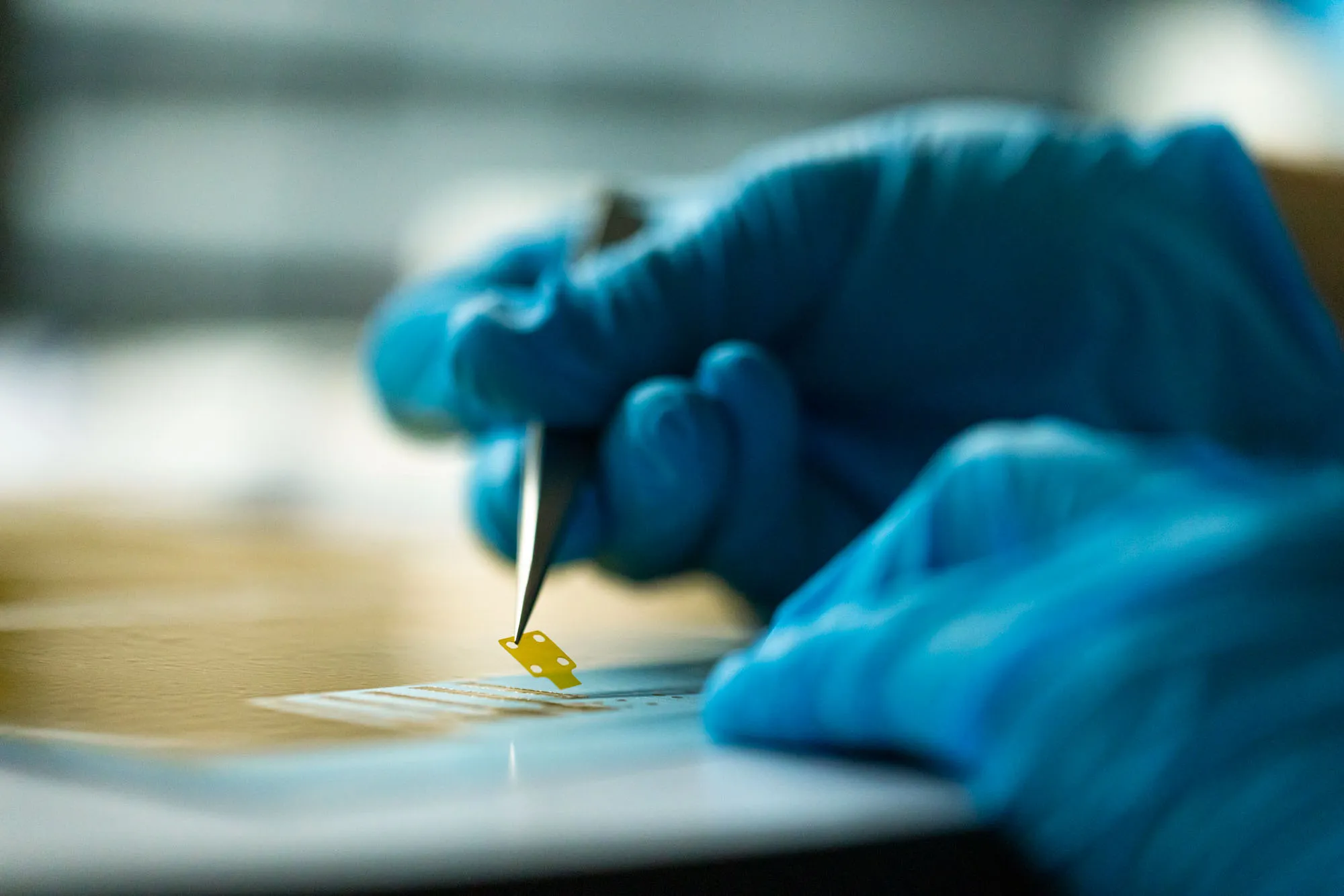 A photo of a scientist wearing gloves using tweezers to pick up a thin film device.