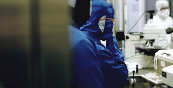 Operators in bunny suits and goggles are seen working around microscopes.